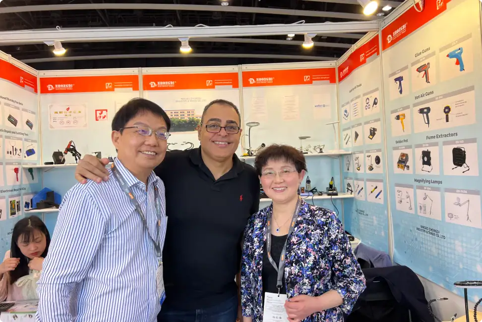 A group of three smiling professionals at an electronics trade show booth showcasing various tools and devices.
