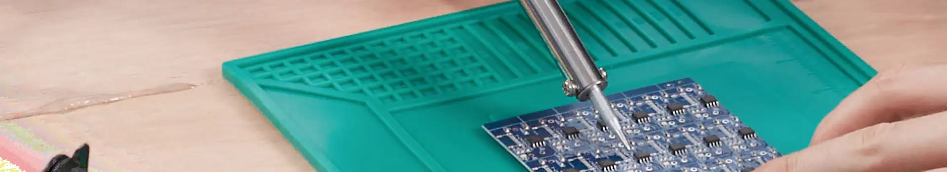 An image showing precise soldering of electronic components onto a circuit board using a soldering iron. The workspace includes a green anti-static mat and a hand carefully working on the electronics.