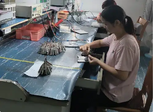 A woman working on electronic components at a workbench with testing equipment and wires, focusing on assembling or inspecting circuit boards in a professional electronics lab.