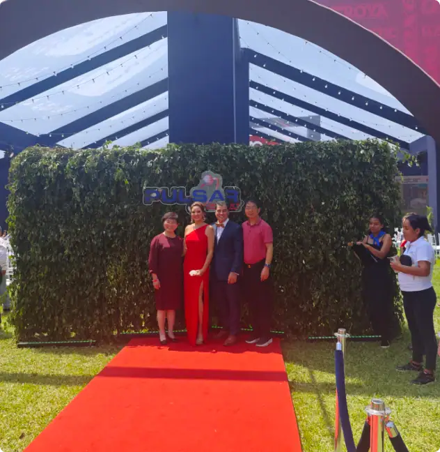 A group of five people standing on a red carpet in front of a green hedge with a Pulsar sign, at an outdoor event. Two women and three men, some in formal attire, posing for a photo with event staff c.