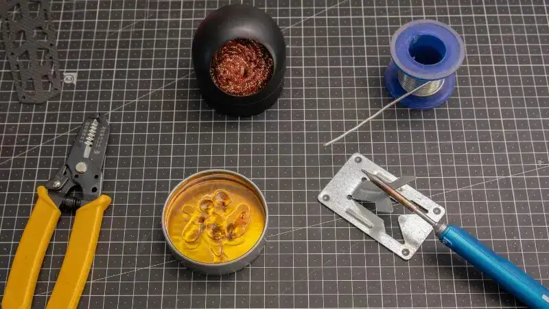 Tools and materials for tinning a soldering iron, including a sponge, solder wire, and a soldering iron stand, on a workbench.