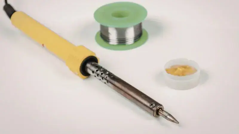 Close-up of a soldering iron with a yellow handle, a spool of solder wire, and a small container of flux, used for electronics soldering.